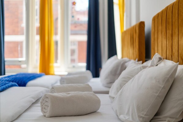 A neatly made bed with white pillows and a rolled white towel, a wooden headboard, and a view of a cityscape through large windows with yellow curtains.