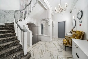 A grand staircase with marble steps leading to a landing with a chandelier and a seating area with a yellow chair.