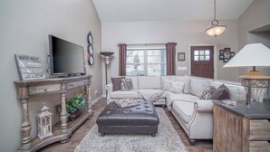 A living room with a large flat-screen TV, a sofa, an ottoman, a lamp, and a decorative sign. - New! Charming Home in the Heart of Bemus Point, Lake Chautauqua, NY (Bemus Point)