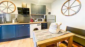 Een compacte keuken met blauwe kastjes, een magnetron, een wasmachine en een eettafel met een gestreepte tafelkleed.