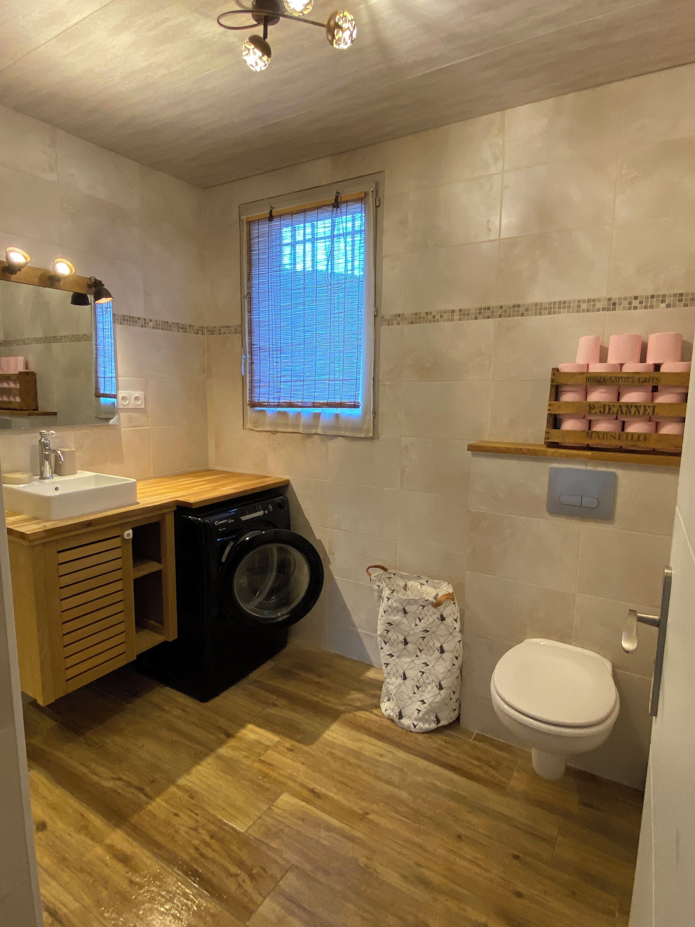 A compact bathroom with a washing machine, a toilet, a sink, and a window with blinds.