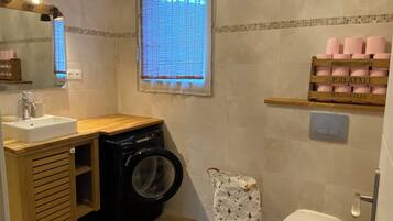 A compact bathroom with a washing machine, a toilet, a sink, and a window with blinds.