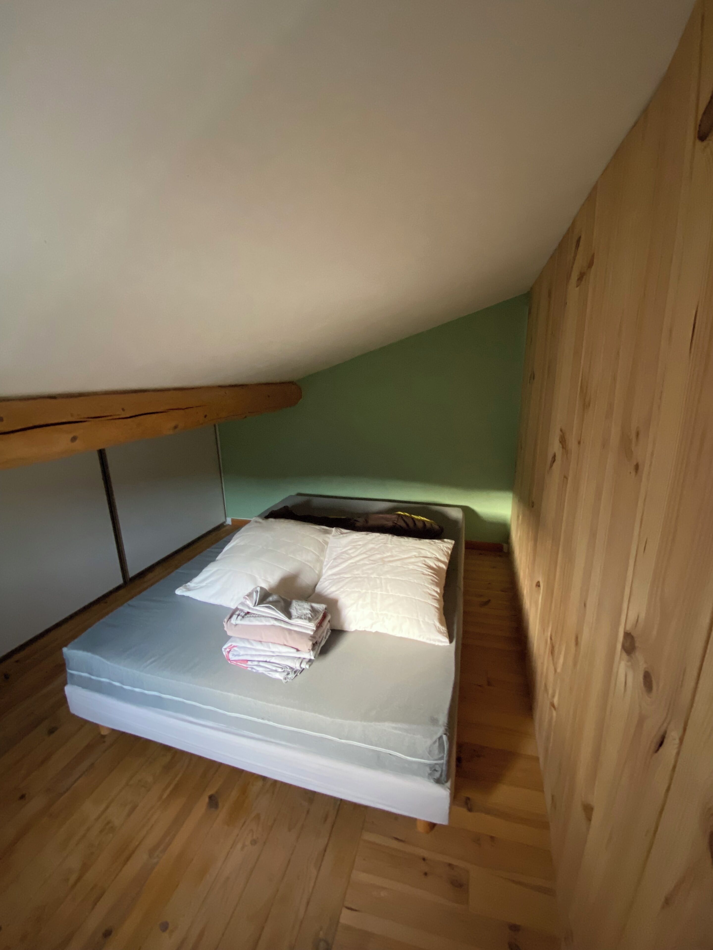 A small, single-bed room with a wooden ceiling and walls, a mattress, pillows, and folded clothes on the bed.