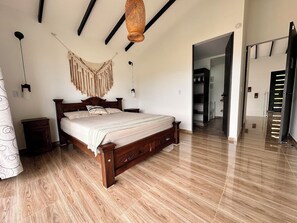 A bedroom with a wooden bed, a hanging macrame wall decoration, and a hallway leading to a bathroom.