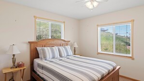 Une chambre à coucher avec un lit en bois, deux tables de chevet, un ventilateur de plafond et deux fenêtres équipées de stores.