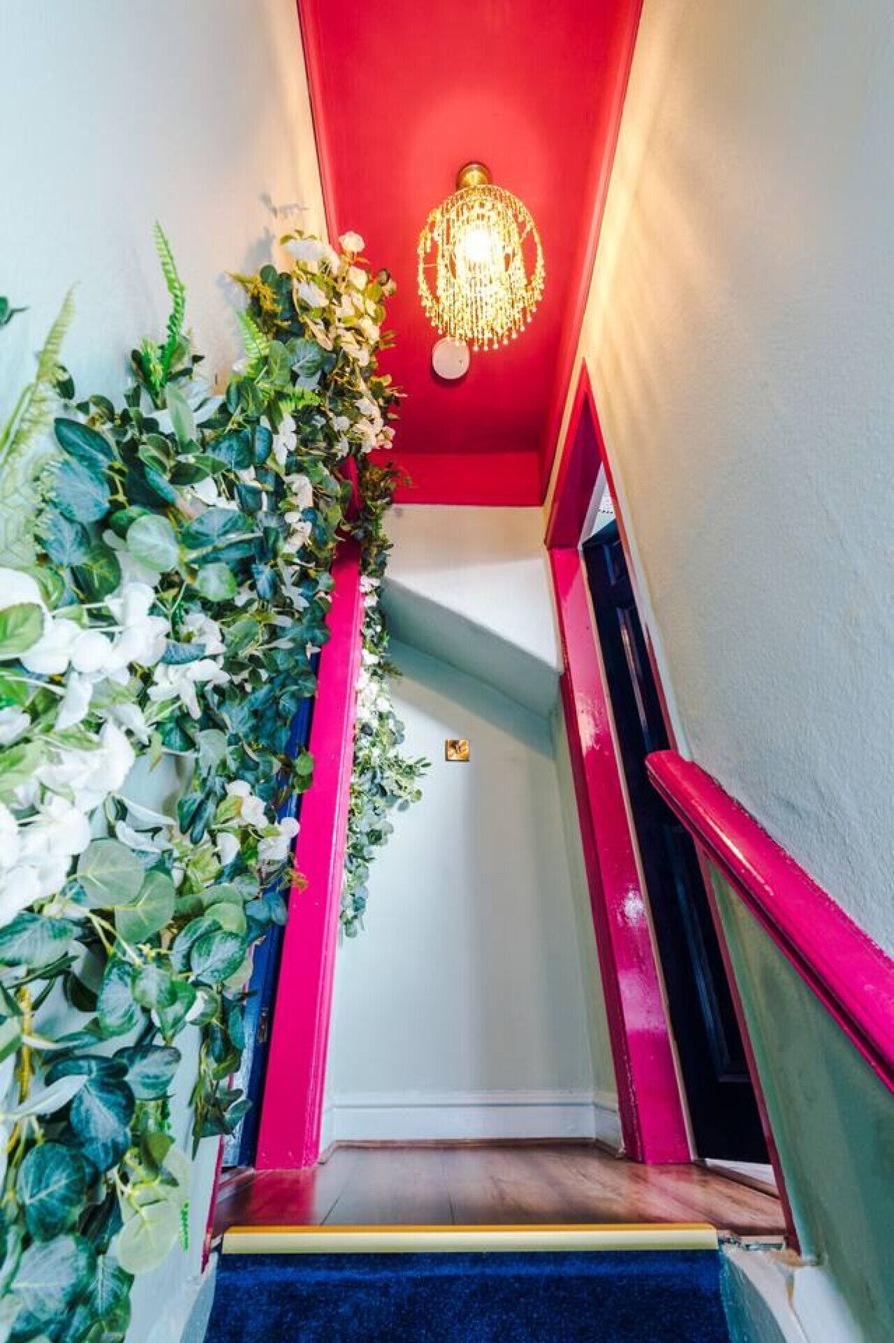 Una escalera con barandilla roja, un candelabro y una pared decorada con follaje verde y flores blancas.