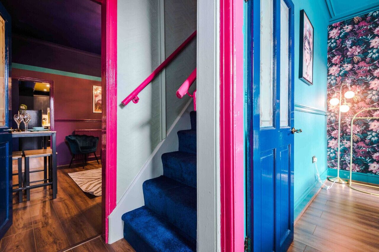A staircase with blue steps and a pink handrail, leading to a room with a floral wallpaper and a chandelier.