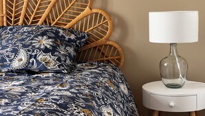 A bed with a patterned duvet and pillows, a wicker headboard, and a bedside table with a lamp and a glass bottle.