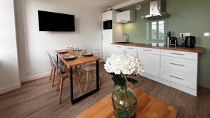 A modern kitchen with a wooden dining table set for four, a large vase with white flowers, and a flat-screen TV mounted on the wall.
