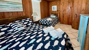 A room with wooden walls and ceiling, two beds with whale-patterned bedding, a ceiling fan, and a window with blinds.
