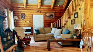 A cozy log cabin interior with a wooden staircase, a beige sofa, a wooden coffee table, and a television in a corner.