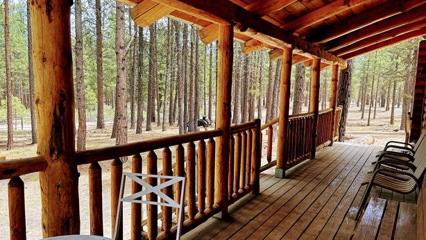 A wooden deck with a railing, a table, and chairs, overlooking a forested area.