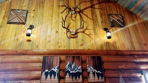 A wooden ceiling with a mounted deer head and two wall-mounted lanterns. Below, there are three wooden panels with mountain designs.