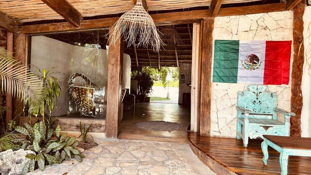 Un espacio al aire libre techado con suelo de piedra, vigas de madera, una lámpara colgante, una bandera y un banco rústico.