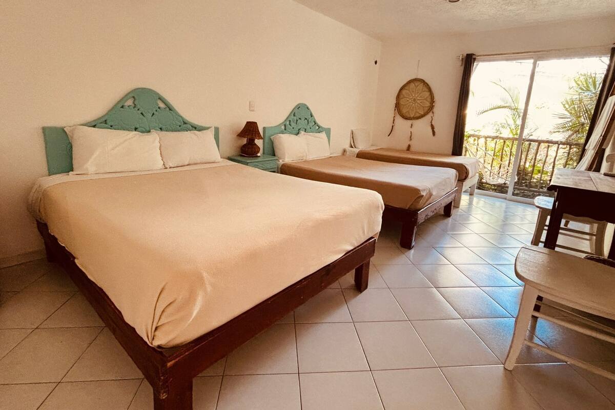 A hotel room with two beds, a small table, and a balcony with a view of palm trees.