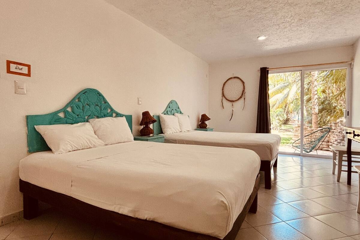 A hotel room with two beds, a small table, a dreamcatcher, and a view of palm trees through the door.