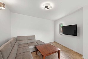 Un salón con un sofá seccional, una mesa de centro de madera, un televisor de pantalla plana montado en la pared y una lámpara en el techo.