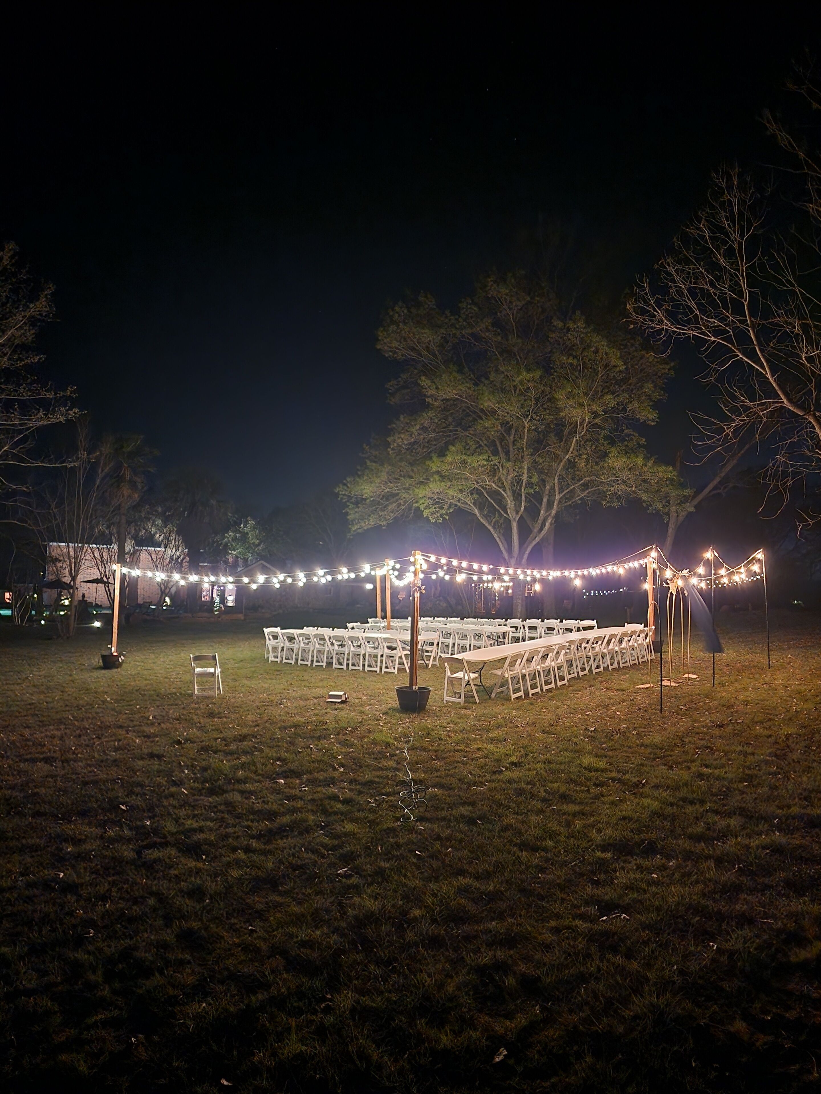 An outdoor setting with rows of white chairs arranged for an event, illuminated by string lights.
