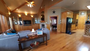 A spacious living room with a large grey sofa, wooden ceiling beams, a ceiling fan, and a kitchen area with wooden cabinets and a refrigerator.