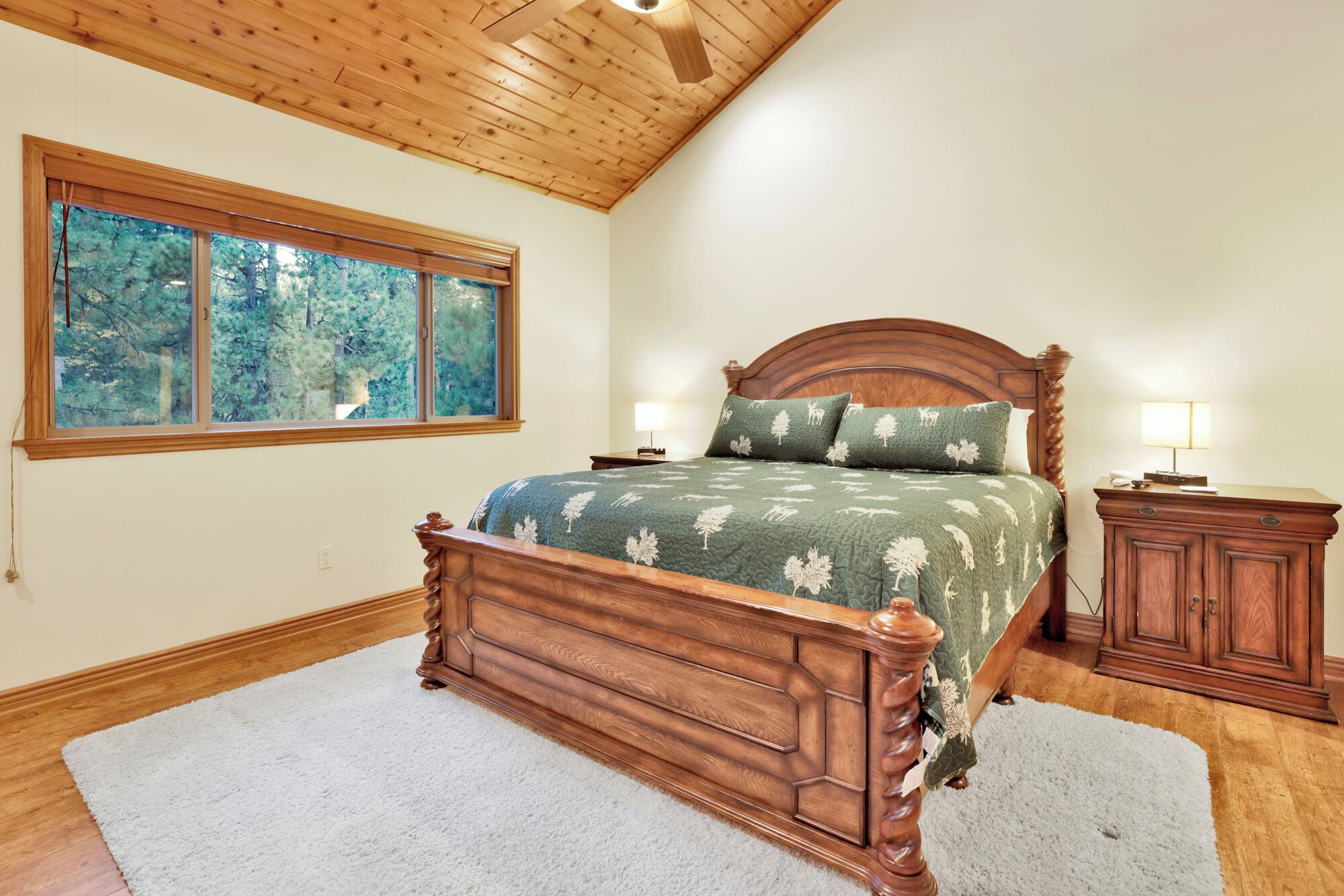 Um quarto com uma cama de madeira, uma janela grande com vista para árvores, uma mesa de cabeceira e um armário.