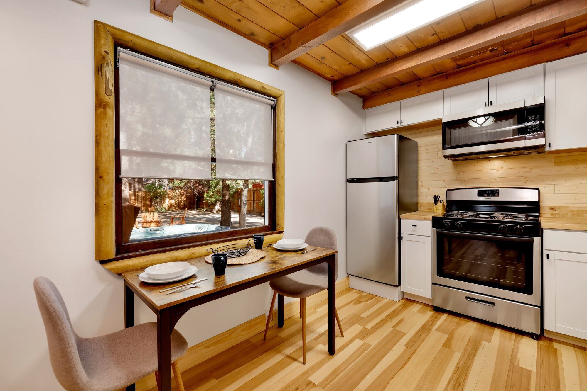 A compact kitchen with a dining area, featuring a wooden ceiling, white cabinets, and a view through a window.
