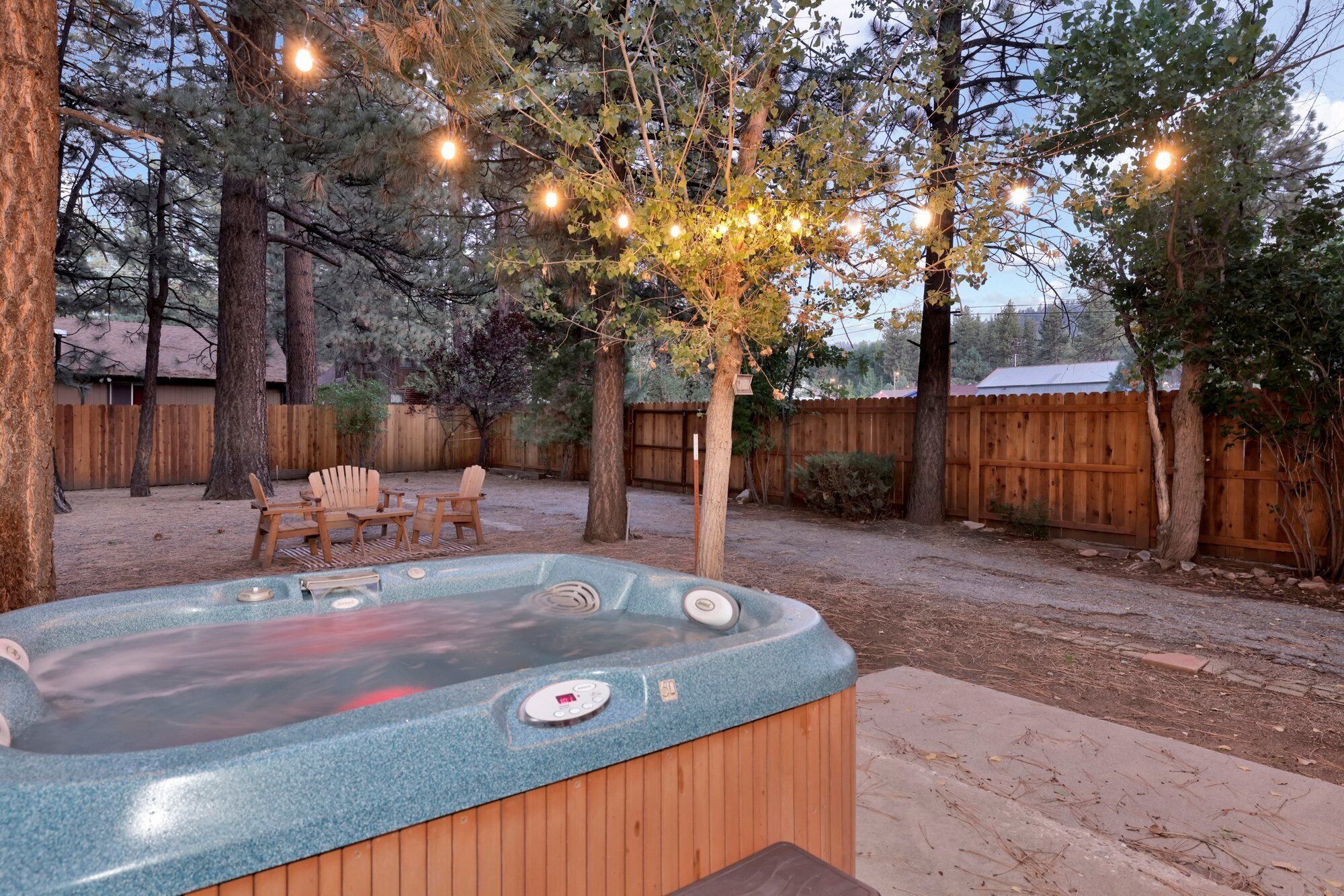 Un jacuzzi en un patio trasero con valla de madera, árboles y luces colgantes.