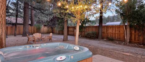 Un jacuzzi en un patio trasero con valla de madera, árboles y luces colgantes.