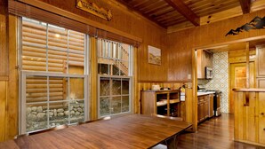 A wooden cabin interior with a dining table, kitchen area, and large windows with blinds. - Silvertip Pines #2335 (Big Bear Lake)