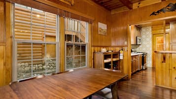 A wooden cabin interior with a dining table, kitchen area, and large windows with blinds.