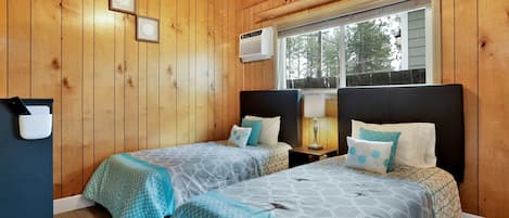 A cabin-style bedroom with two beds, wooden walls, a window with a view, and a mounted air conditioning unit.