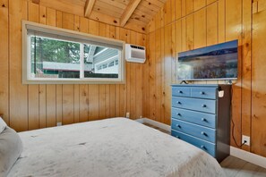 Een kamer met houten lambrisering, een bed, een dressoir, een kachel en een raam met uitzicht op een huis en bomen.