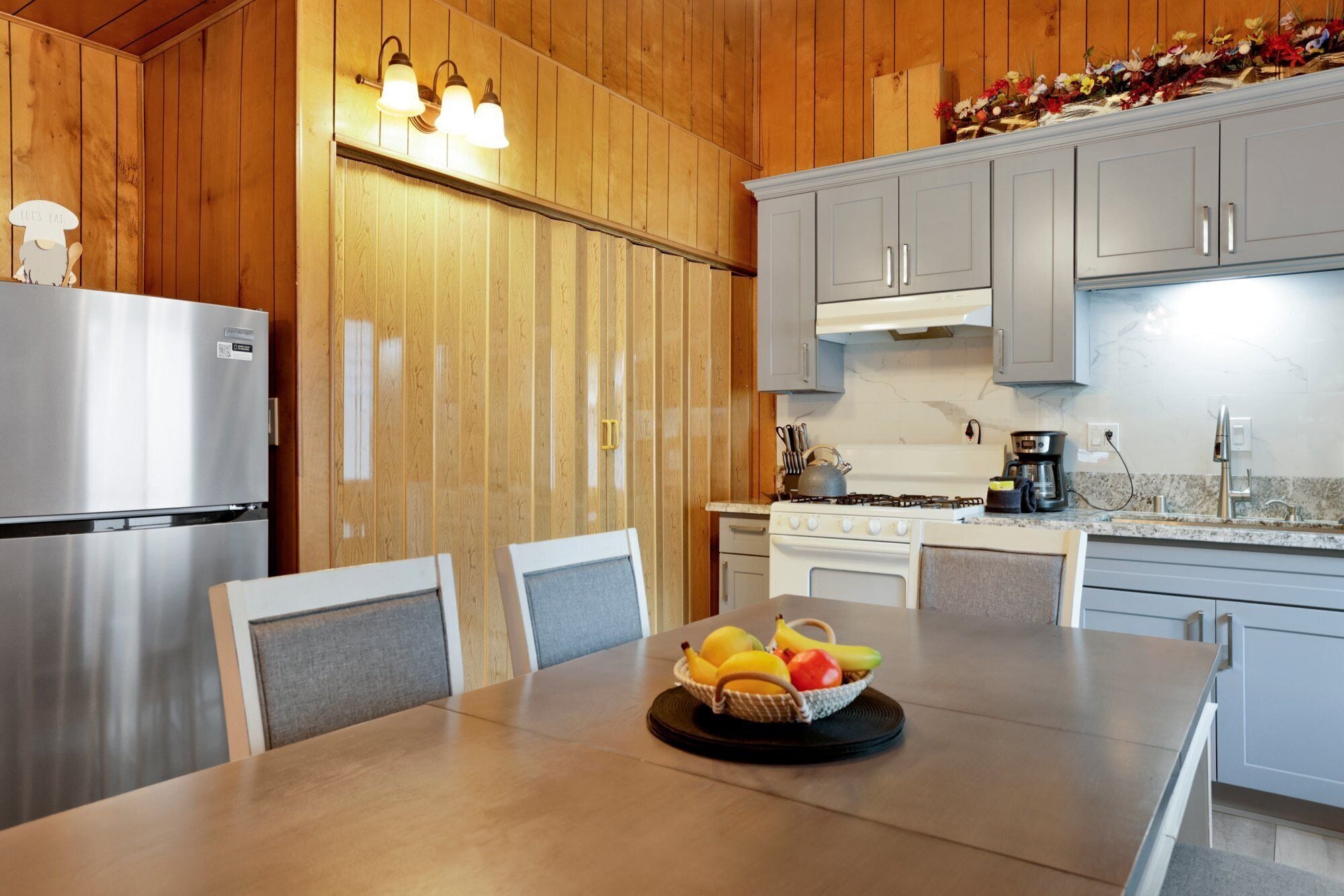 A modern kitchen with wooden paneling, a stainless steel refrigerator, a stove, and a dining table with a fruit basket.