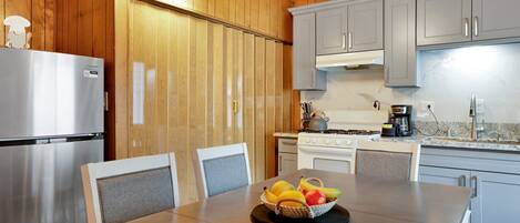 A modern kitchen with wooden paneling, a stainless steel refrigerator, a stove, and a dining table with a fruit basket.