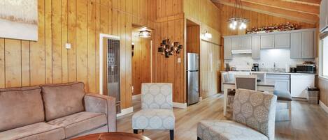 A spacious living area with a wooden ceiling, a sofa, two armchairs, and a coffee table. A kitchen is visible in the background with modern appliances and cabinets.
