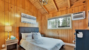 A wooden cabin bedroom with a bed, bedside table, lamp, and a window with a view of trees.