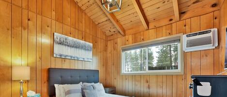 Dormitorio de una cabaña de madera con cama, mesita de noche, lámpara y ventana con vistas a árboles.