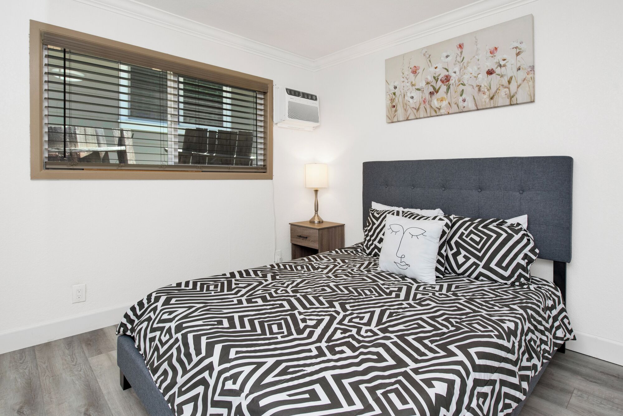 A bedroom with a bed, a nightstand, a lamp, a window with blinds, and a floral artwork on the wall.