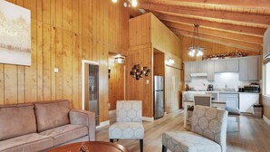 Un vaste espace de vie doté d’un plafond en bois, d’un canapé, de deux fauteuils et d’une table basse. On aperçoit une cuisine à l’arrière-plan, équipée d’appareils électroménagers et de meubles de rangement modernes.