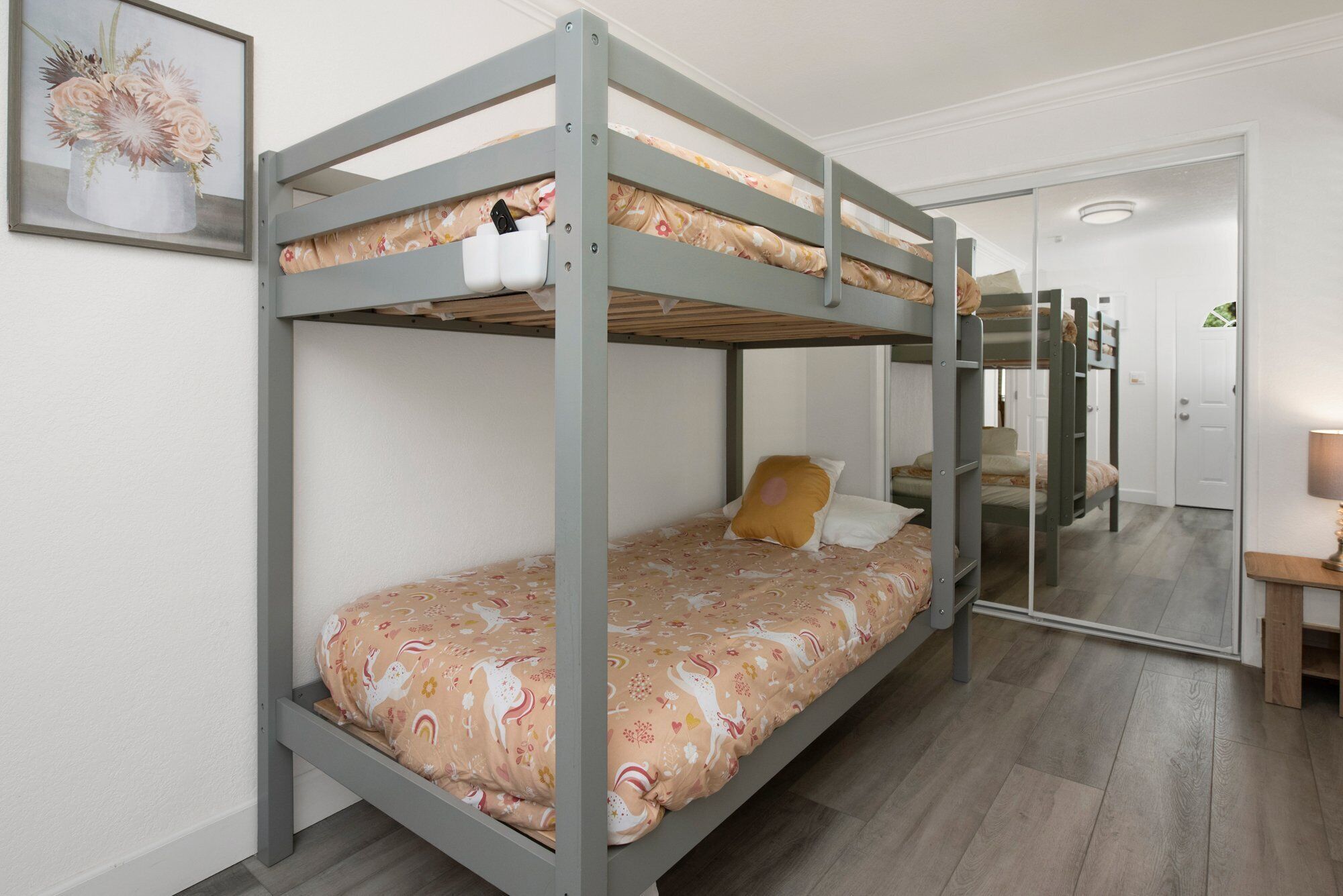 A bunk bed room with a floral bedspread, a small table with a lamp, and a mirror on the wall.