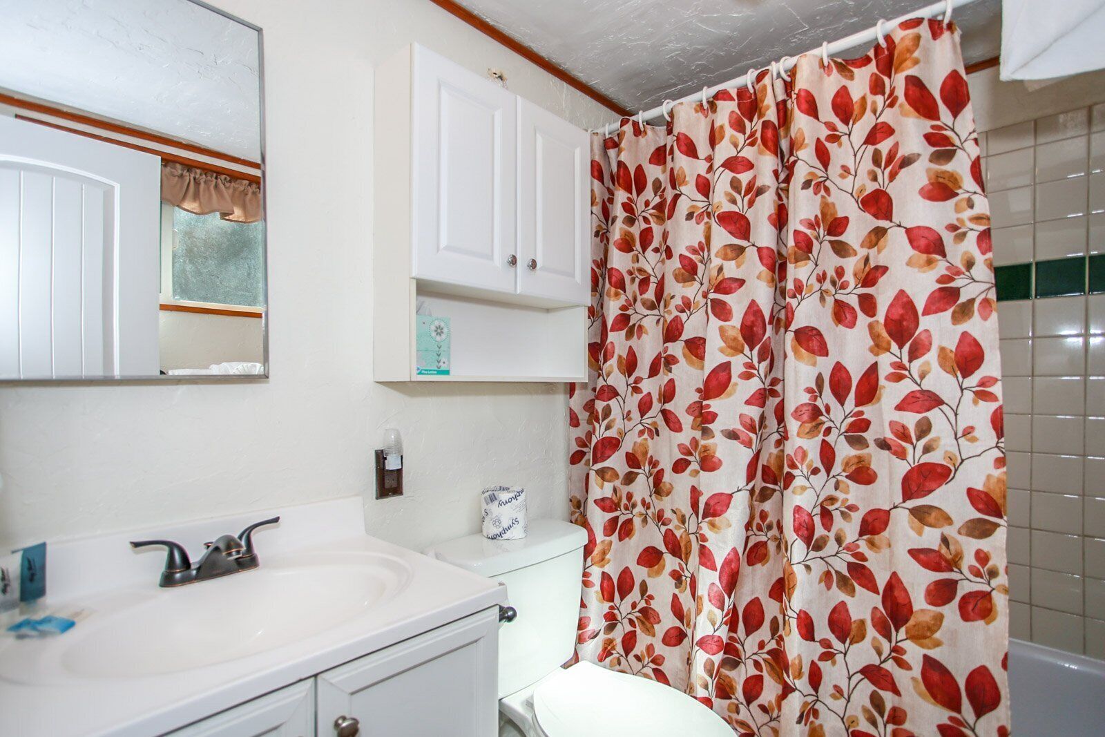 A bathroom with a white sink, mirror, and cabinet, a toilet, and a shower curtain with a leaf pattern.