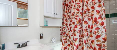 A bathroom with a white sink, mirror, and cabinet, a toilet, and a shower curtain with a leaf pattern.