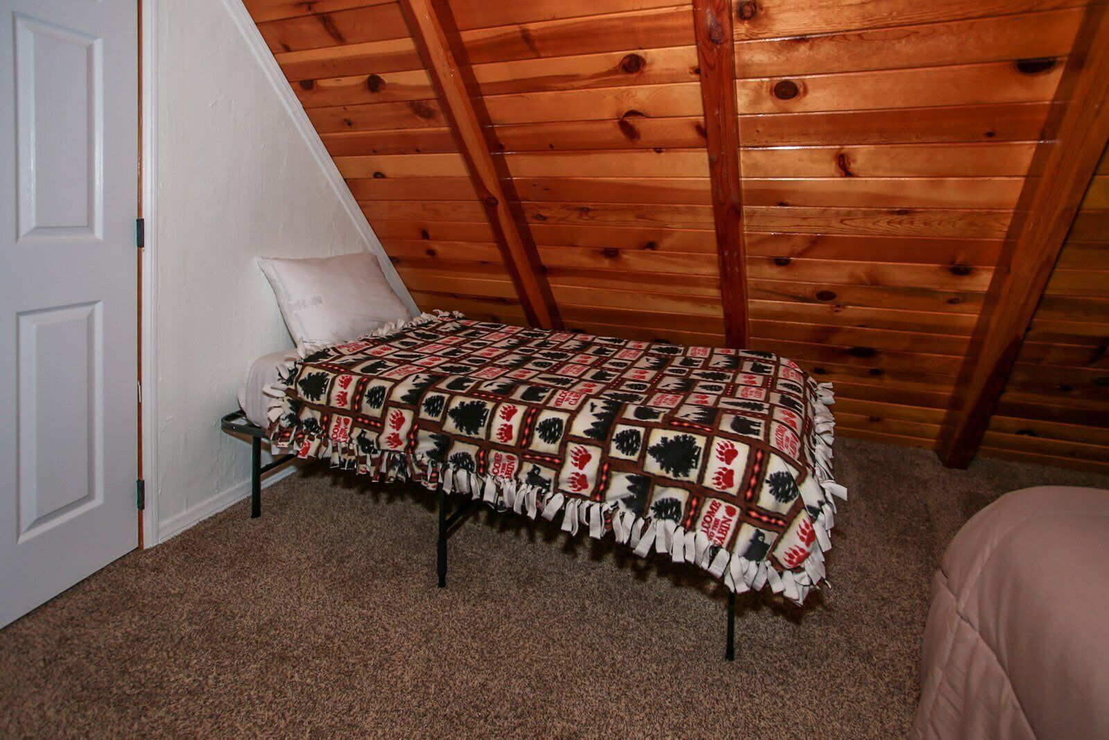 A small, cozy room with a sloped ceiling and wooden beams. A single bed with a white headboard is positioned against a white door.