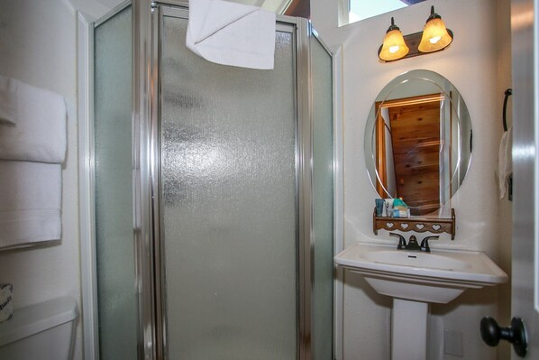 Une salle de bain avec une porte de douche en verre, un lavabo blanc, un miroir avec un cadre en bois et deux appliques murales.
