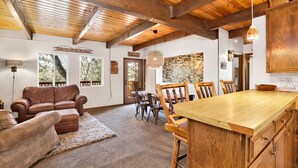 A cozy interior with a wooden ceiling, a dining area with a wooden table and chairs, a leather sofa, and a rug. - Chipmunk Treehouse #1983 (Big Bear Lake)