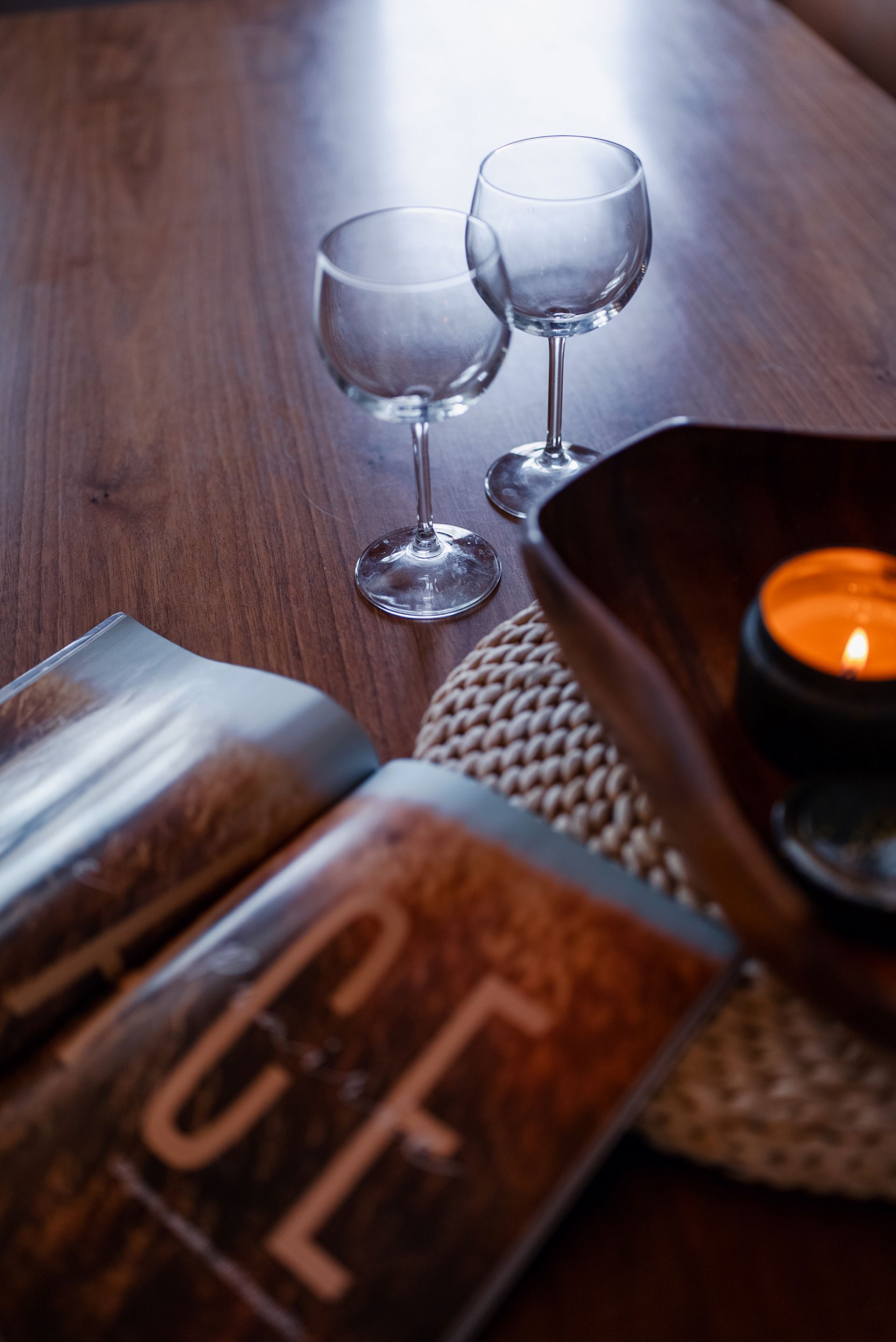 A wooden table with two glasses and a candle, a book, and a woven placemat.