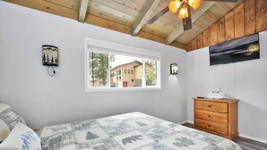 A bedroom with a bed, a wooden dresser, a window with a view of a house, and wall-mounted lights.