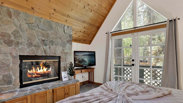 A bedroom with a stone fireplace, a television, and a large window with a view of trees. - Green Canoe Cabin #2293 (Big Bear Lake)