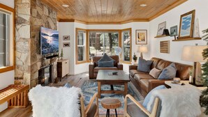 A cozy living room with a stone fireplace, wooden ceiling, and large windows offering a view of the outdoors. - Green Canoe Cabin #2293 (Big Bear Lake)