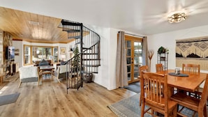 A spacious living area with a wooden floor, a fireplace, a dining table, and a spiral staircase. - Green Canoe Cabin #2293 (Big Bear Lake)