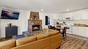 Una sala de estar con chimenea de piedra, un sofá de cuero, un comedor con mesa y sillas, y una cocina con armarios y electrodomésticos blancos.
